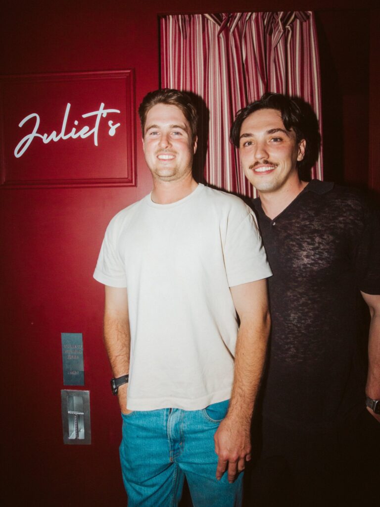 Two men smiling in front of the red Juliet's Photobooth sign and striped curtain on Brunswick Street, New Farm